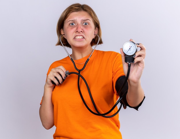 Jovem insalubre em uma camiseta laranja se sentindo mal, medindo a pressão arterial usando o tonômetro, parecendo preocupada e desapontada em pé sobre uma parede branca