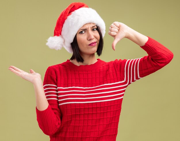 Jovem insatisfeita com chapéu de Papai Noel, olhando para a câmera, mostrando a mão vazia e o polegar para baixo, isolado em um fundo verde oliva