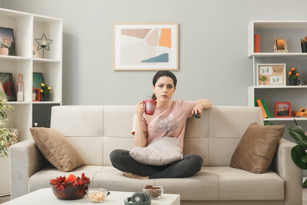 Foto grátis jovem insatisfeita segurando o controle remoto da tv com uma xícara de chá, sentada no sofá atrás da mesa de centro da sala de estar
