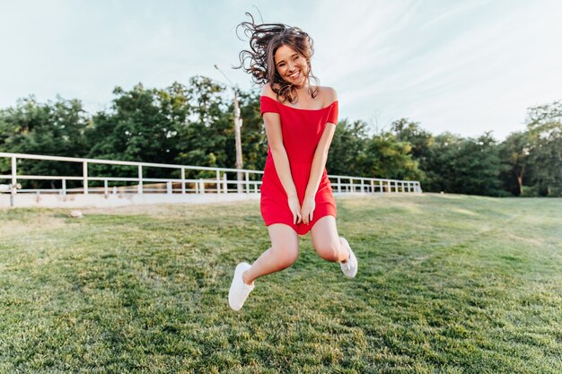 Jovem inspirada pulando no parque e sorrindo. Linda menina morena com vestido vermelho, se divertindo no fim de semana de verão.
