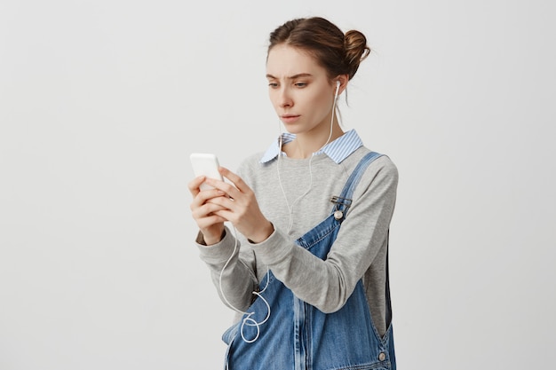 Jovem intrigada em fones de ouvido franzindo a testa tendo problema com o gadget. linda produtora que está decepcionada por causa da falta de conexão à internet em seu telefone. conceito de tecnologia