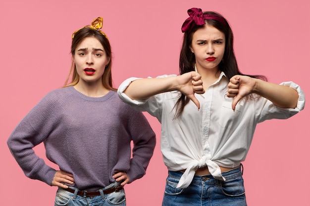 Jovem irritada com a testa franzida mantendo as mãos na cintura sendo teimosa posando ao lado de sua amiga irritada e irritada que está mostrando o polegar para baixo gesto de antipatia como sinal de desaprovação