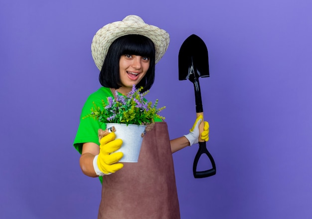 Jovem jardineira alegre de uniforme, usando chapéu de jardinagem, segurando um vaso de flores e uma pá