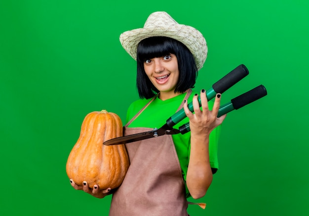 Jovem jardineira alegre de uniforme, usando chapéu de jardinagem, segurando uma abóbora e uma tesoura