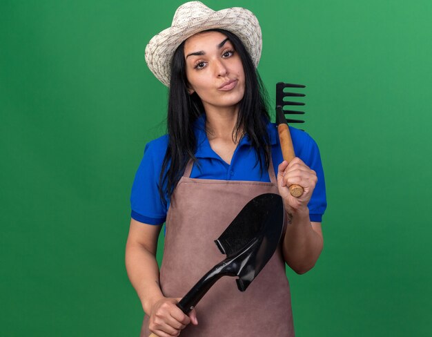 Jovem jardineira caucasiana confiante usando uniforme e chapéu segurando o ancinho no ombro e a pá
