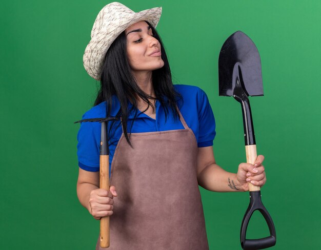 Jovem jardineira caucasiana satisfeita usando uniforme e chapéu segurando o ancinho e a pá olhando para a pá