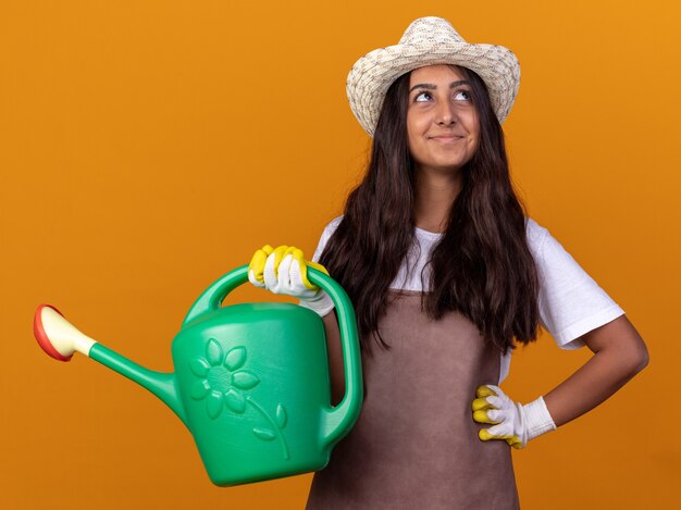 Jovem jardineira com avental e chapéu de verão segurando um regador, olhando para cima com um sorriso no rosto em pé sobre a parede laranja