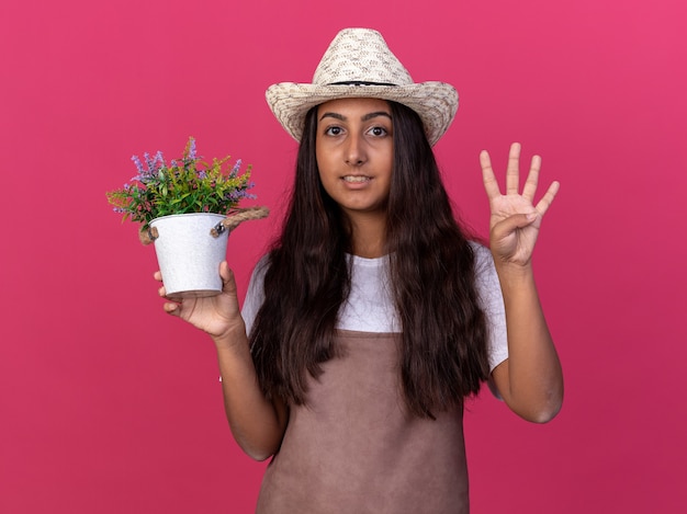 Foto grátis jovem jardineira com avental e chapéu de verão segurando um vaso de planta sorrindo e mostrando o número quatro em pé sobre a parede rosa