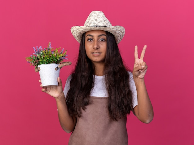 Jovem jardineira com avental e chapéu de verão segurando um vaso de planta sorrindo e mostrando o sinal v em pé sobre a parede rosa