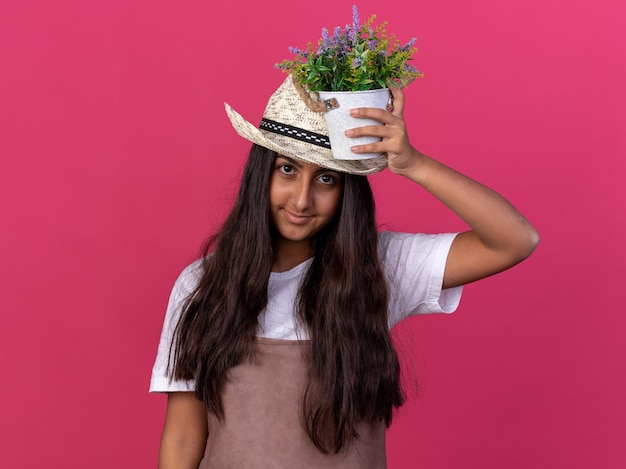 Foto grátis jovem jardineira com avental e chapéu de verão segurando um vaso de plantas na cabeça com um sorriso no rosto em pé sobre a parede rosa