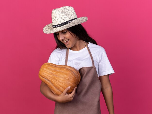 Jovem jardineira com avental e chapéu de verão segurando uma abóbora olhando para ela com um sorriso no rosto feliz e animada em pé sobre a parede rosa