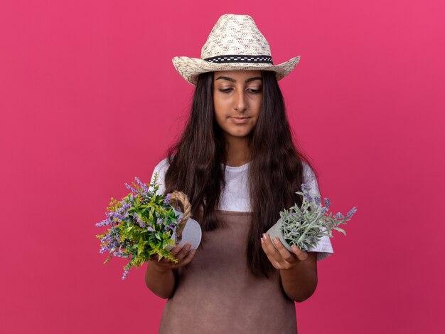 Jovem jardineira com avental e chapéu de verão segurando vasos de plantas olhando para eles com uma cara séria em pé sobre a parede rosa