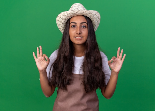 Jovem jardineira com avental e chapéu de verão sorrindo mostrando sinal de ok com as duas mãos em pé sobre a parede verde