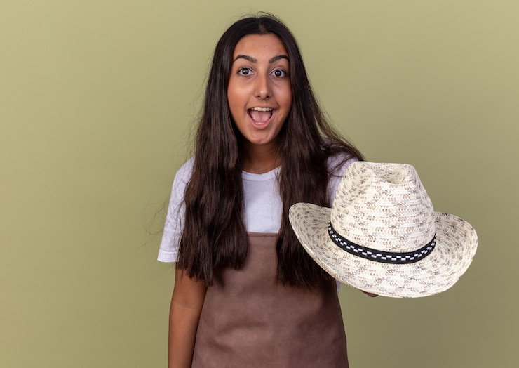 Foto grátis jovem jardineira com avental mostrando seu chapéu de verão feliz e surpresa sorrindo em pé sobre a parede verde