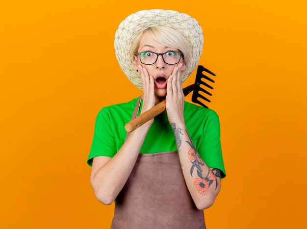 Foto grátis jovem jardineira com cabelo curto, avental e chapéu segurando um mini rake olhando para a câmera espantada e surpresa em pé sobre um fundo laranja