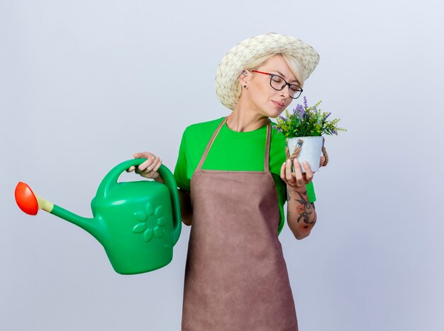 Jovem jardineira com cabelo curto no avental e chapéu segurando um regador e um vaso de planta sentindo um aroma agradável