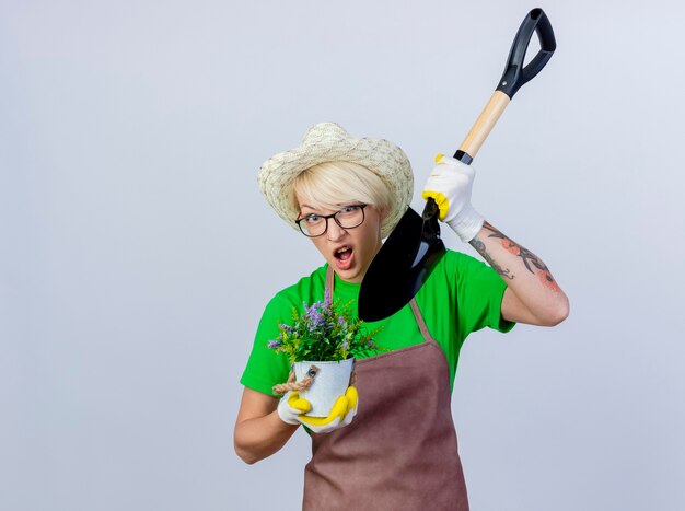 Jovem jardineira com cabelo curto no avental e chapéu segurando uma pá e um vaso de planta sendo confundida
