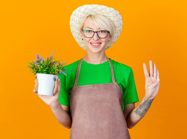 Jovem jardineira com cabelo curto no avental e chapéu segurando uma planta em um vaso, olhando para a câmera, sorrindo com uma cara feliz, mostrando o número quatro em pé sobre um fundo laranja