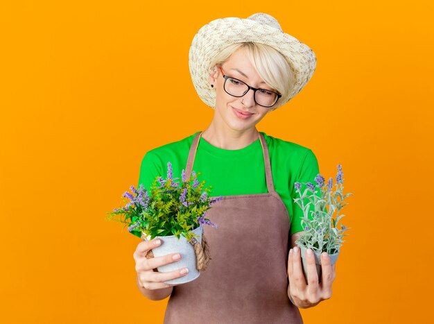 Jovem jardineira com cabelo curto no avental e chapéu segurando vasos de plantas olhando para eles sorrindo com uma cara feliz em pé sobre um fundo laranja