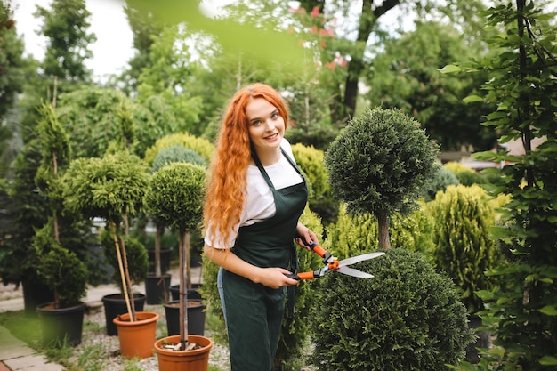 Jovem jardineira com cabelo encaracolado ruivo em pé no avental e segurando uma tesoura de jardim grande enquanto alegremente olhando na câmera ao ar livre