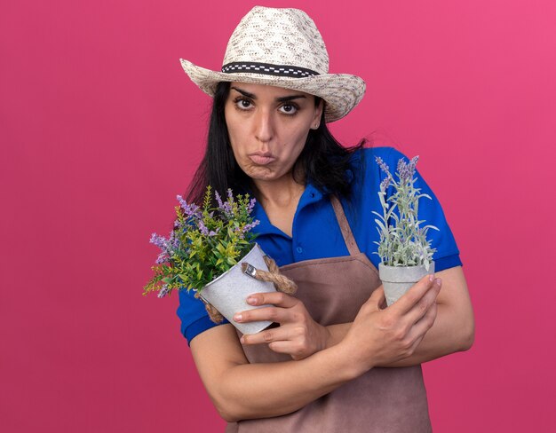 Jovem jardineira confusa, usando uniforme e chapéu, segurando vasos de flores, olhando para a frente, isolada na parede rosa com espaço de cópia