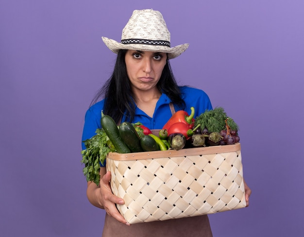 Jovem jardineira confusa vestindo uniforme e chapéu segurando uma cesta de legumes, olhando para a frente, isolada na parede roxa com espaço de cópia
