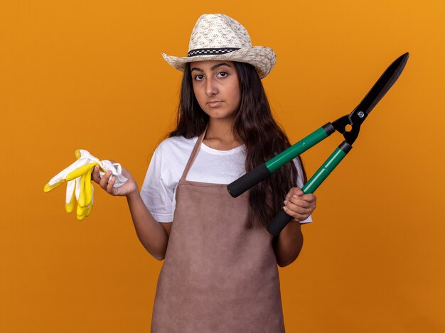 Jovem jardineira de avental e chapéu de verão segurando uma tesoura de sebe e luvas de trabalho com um sorriso no rosto em pé sobre uma parede laranja