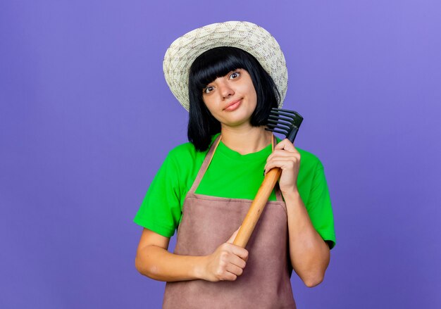 Jovem jardineira empolgada de uniforme usando chapéu de jardinagem segurando o ancinho