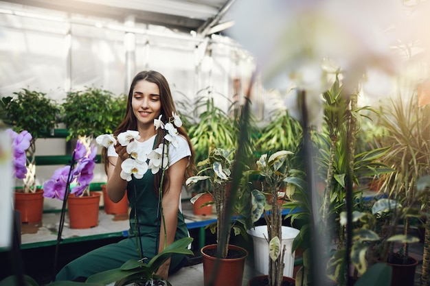 Jovem jardineira fazendo uma pausa em seu trabalho sentada na estufa conversando com flores