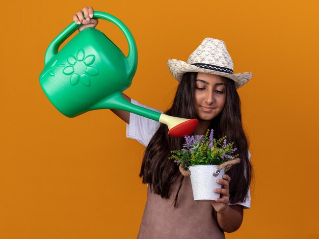 Jovem jardineira feliz com avental e chapéu de verão segurando um regador e um vaso de planta regadora com um sorriso no rosto em pé sobre a parede laranja