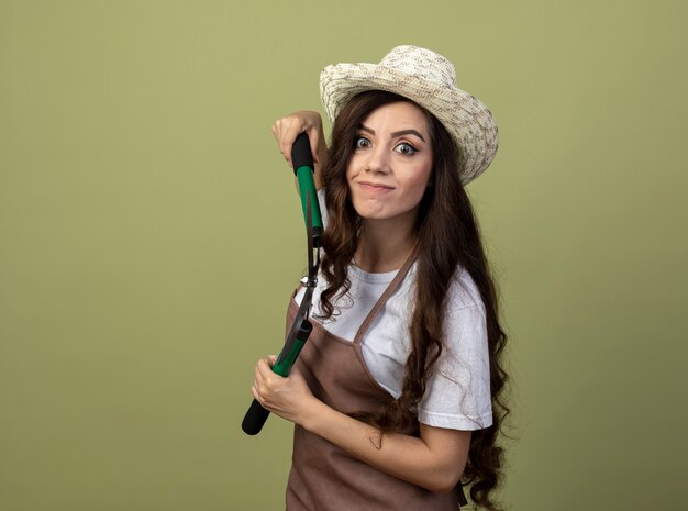 Jovem jardineira impressionada de uniforme, usando chapéu de jardinagem, parada de lado segurando uma tesoura de jardim isolada na parede verde oliva