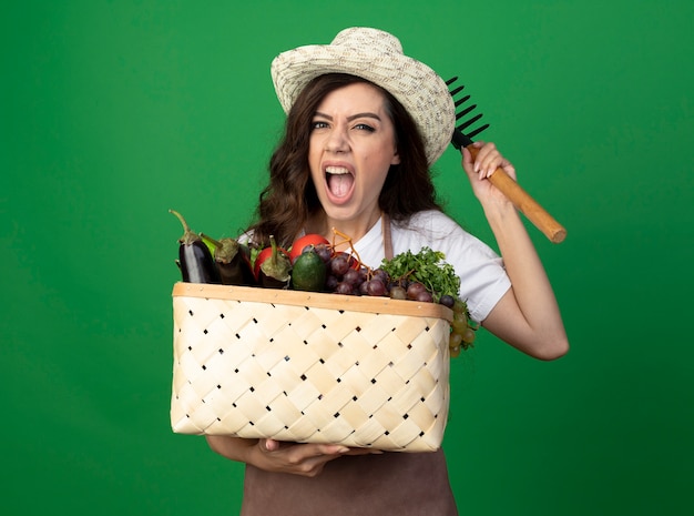 Jovem jardineira irritada de uniforme, usando chapéu de jardinagem, segurando uma cesta de vegetais e um ancinho isolados na parede verde