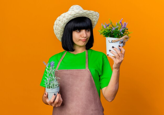 Jovem jardineira pensativa de uniforme, usando um chapéu de jardinagem, segurando e olhando para vasos de flores