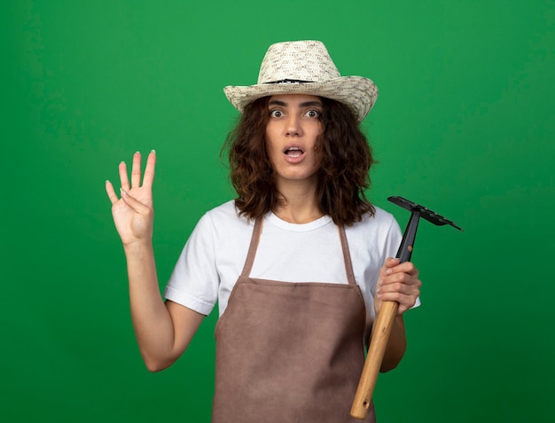 Jovem jardineira preocupada de uniforme, usando chapéu de jardinagem, segurando o ancinho, mostrando quatro