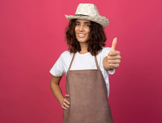 Jovem jardineira sorridente de uniforme usando chapéu de jardinagem, colocando a mão no quadril e mostrando o polegar