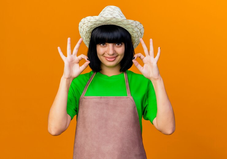 Foto grátis jovem jardineira sorridente de uniforme usando chapéu de jardinagem gesticulando com a mão bem sinalizada