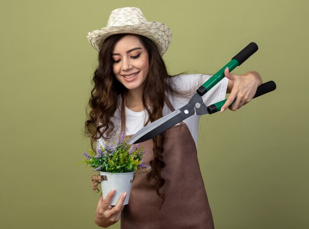 Jovem jardineira sorridente de uniforme usando chapéu de jardinagem segura a tesoura de jardim sobre flores em um vaso de flores isolado na parede verde oliva