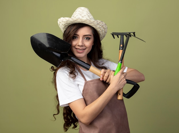 Jovem jardineira sorridente de uniforme usando chapéu de jardinagem segura ferramentas de jardinagem isoladas na parede verde oliva