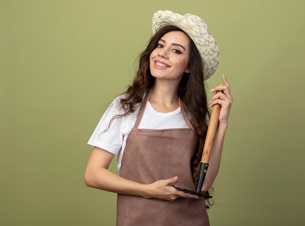 Jovem jardineira sorridente de uniforme usando chapéu de jardinagem segura o ancinho de cabeça para baixo isolado na parede verde oliva