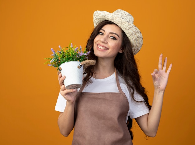 Jovem jardineira sorridente de uniforme usando chapéu de jardinagem segura o vaso de flores e gesticula sinal de mão ok isolado na parede laranja com espaço de cópia