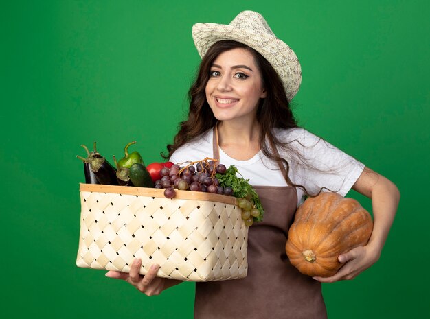 Jovem jardineira sorridente de uniforme usando chapéu de jardinagem segura uma cesta de vegetais e uma abóbora isolada em uma parede verde com espaço de cópia