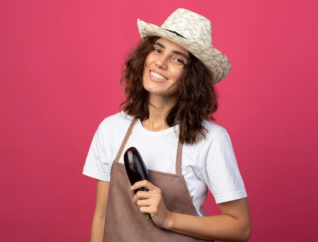 Jovem jardineira sorridente de uniforme usando chapéu de jardinagem segurando berinjela