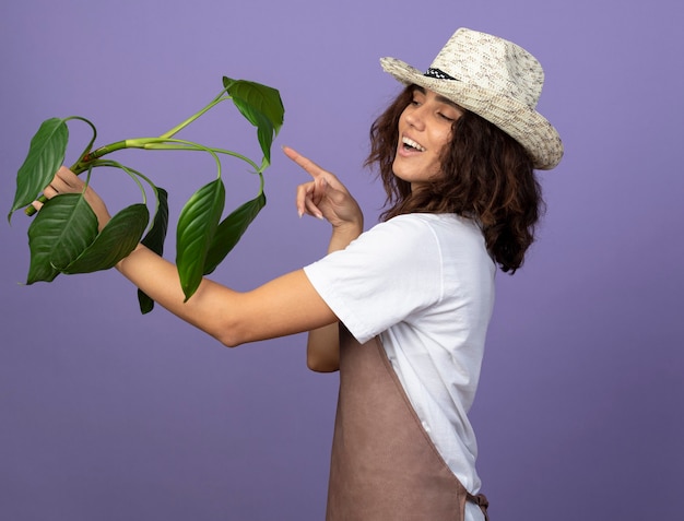 Foto grátis jovem jardineira sorridente de uniforme usando chapéu de jardinagem segurando e apontando para a planta