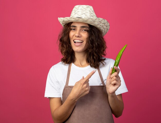 Jovem jardineira sorridente de uniforme usando chapéu de jardinagem segurando e apontando para pimenta isolada em rosa