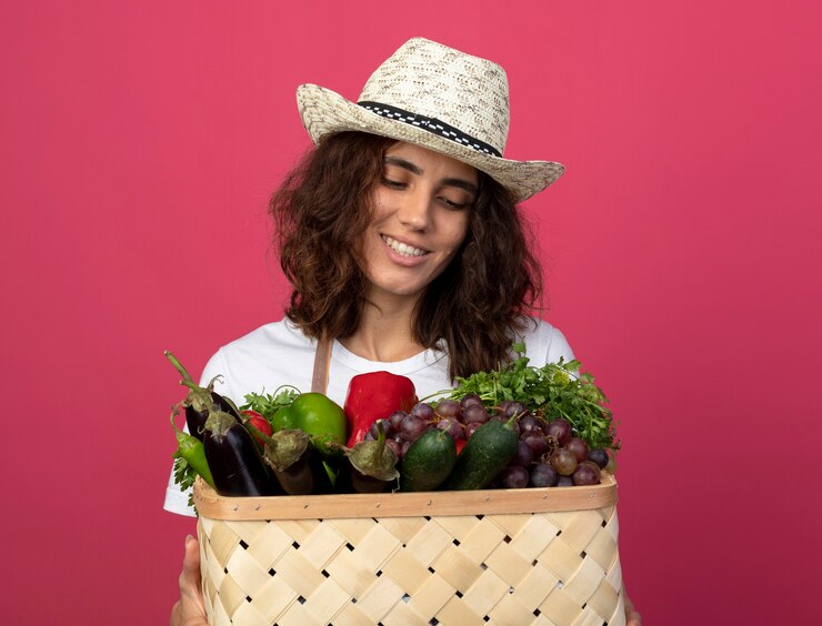 Foto grátis jovem jardineira sorridente de uniforme usando chapéu de jardinagem segurando e olhando para uma cesta de vegetais isolada em rosa