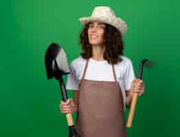 Foto grátis jovem jardineira sorridente de uniforme usando chapéu de jardinagem segurando o ancinho com pá
