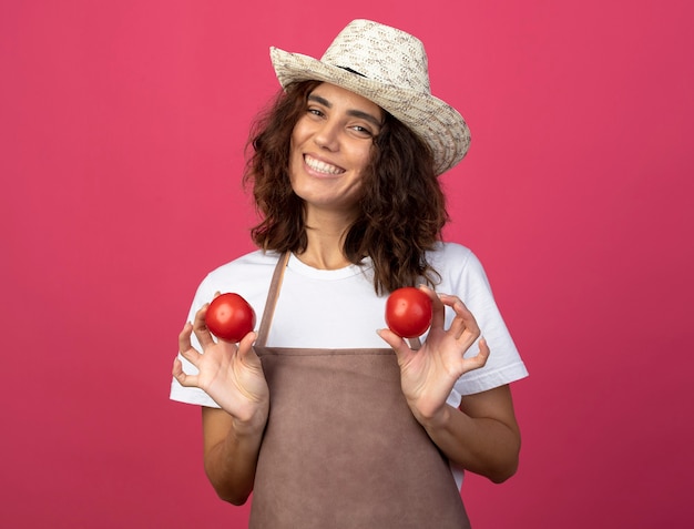 Foto grátis jovem jardineira sorridente de uniforme usando chapéu de jardinagem segurando tomates isolados em rosa