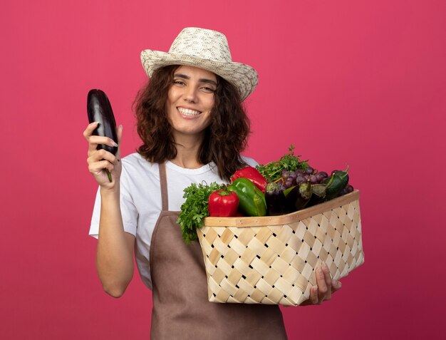 Jovem jardineira sorridente de uniforme usando chapéu de jardinagem segurando uma cesta de vegetais com berinjela