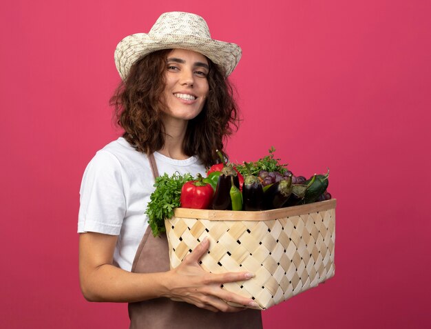 Jovem jardineira sorridente de uniforme usando chapéu de jardinagem segurando uma cesta de vegetais isolada em rosa