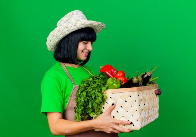 Jovem jardineira sorridente de uniforme usando chapéu de jardinagem segurando uma cesta de vegetais